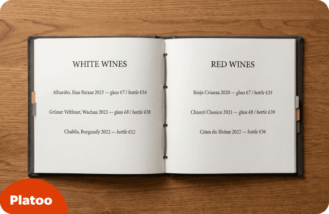 Cuadernillo de carta de vinos abierto en una carpeta oscura de tornillos, con vinos blancos en la página izquierda y tintos en la derecha, sobre una mesa de madera