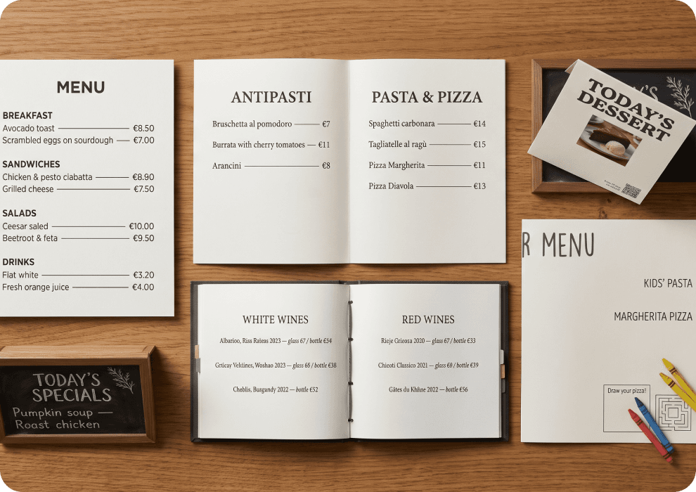 Varios estilos de carta impresa sobre una mesa de madera: carta de una hoja de una cafetería, un díptico abierto, un cuadernillo de carta de vinos, un expositor de mesa, una pizarra y un mantel individual infantil con ceras de colores