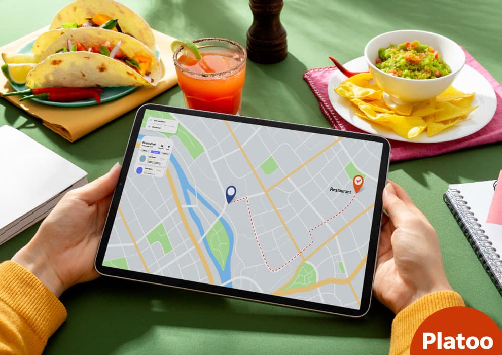 Hands holding a tablet with a map route to a restaurant, next to tacos, guacamole with nachos and a drink on a green table
